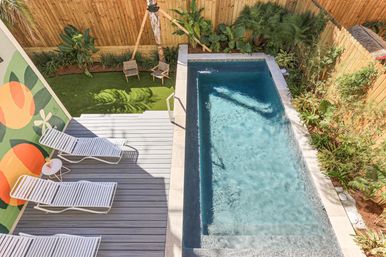Aerial view of a rectangular backyard plunge pool with clear blue water, gray deck with white lounge chairs and small side table, colorful mural on the wall, and tropical plants along a wooden fence.