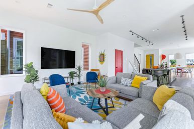Bright modern open-concept living room with gray sectional sofas, colorful throw pillows and rug, wall-mounted TV, blue accent chairs, wood ceiling fan, plants, and kitchen island in the background.