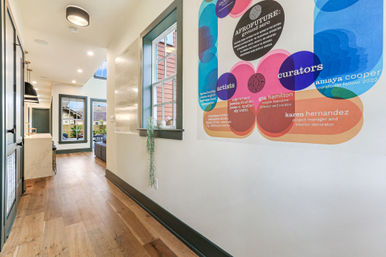 Bright modern townhouse hallway with wide-plank hardwood floors, pendant lights and open kitchen; vibrant colorful exhibition poster on the wall and large windows overlooking a courtyard.