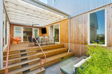 Sunlit covered wooden deck at a contemporary home with wide steps and metal handrail, outdoor dining table and chairs, orange lounge seats, wall-mounted TV, sliding glass doors and a leafy planter.