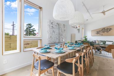 Sunny modern coastal-style open-plan dining and living room with a long wooden table set with turquoise dishes, woven white pendant lights, large windows, light wood floors, and a green sofa — airy, minimalist interior.