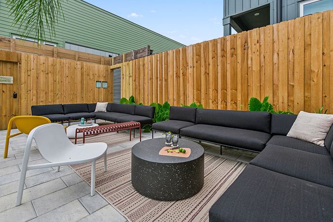 Modern urban patio courtyard with L-shaped black sectional sofa, round speckled stone coffee table, white and yellow accent chairs, patterned rug, concrete pavers, tall wooden privacy fence and greenery.