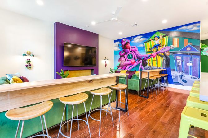 Bright colorful lounge interior with a long wooden bar and round wood stools, green seating, wall-mounted TV, hardwood floors, and a large vibrant mural of a purple octopus musician and neighborhood scene.