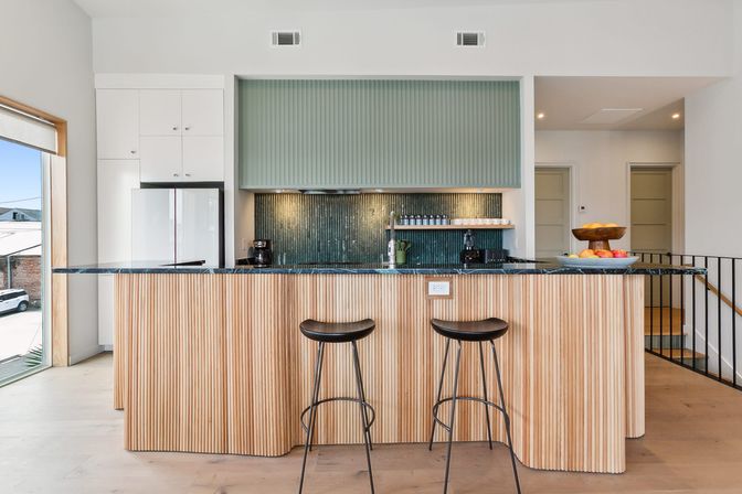 Bright modern open-plan kitchen with fluted wood island and dark marble countertop, two black bar stools, textured green tile backsplash, white cabinets and large sunlit window