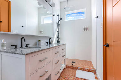 Bright modern bathroom: white double vanity with granite countertop and black faucets, glass walk-in shower with rain and handheld heads, orange penny-tile floor and matching orange door, high window showing blue sky.