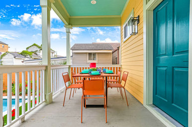 Bright covered porch on a yellow house with a teal front door, orange outdoor dining chairs and table set, suburban backyard view