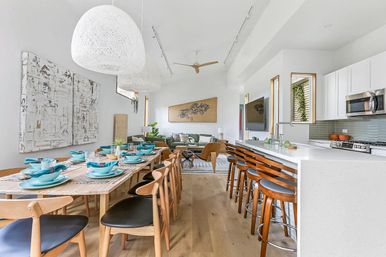 Bright modern open-plan dining and kitchen in a sunlit contemporary home — long wooden table set with turquoise dishes, oversized woven pendant lights, white quartz island with wooden bar stools, hardwood floors and a cozy living area in the background.