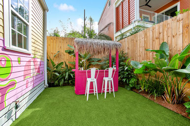Cheerful small backyard with a bright pink tiki bar and thatched roof, two white bar stools on green artificial turf, tropical plants, a wooden fence and colorful mural on the house.
