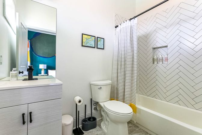 Bright modern residential bathroom with white herringbone tile shower, bathtub with gray-striped curtain with yellow trim, white vanity with black faucet and toilet.
