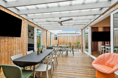 Bright covered wooden deck patio with long black-covered dining table and mixed chairs, ceiling fan, sliding glass doors to indoor living areas, potted plants and a small pool in a fenced private backyard.