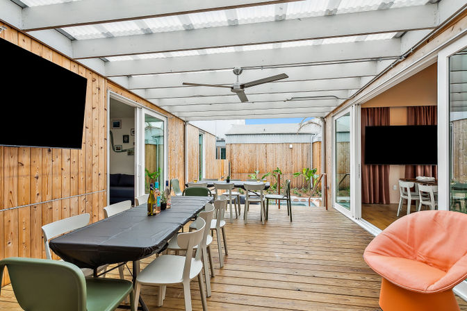 Bright covered wooden deck patio with long black-covered dining table and mixed chairs, ceiling fan, sliding glass doors to indoor living areas, potted plants and a small pool in a fenced private backyard.