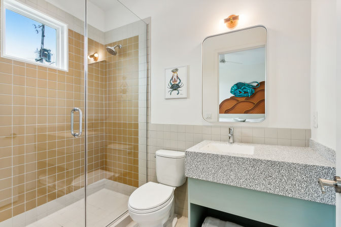 Bright modern bathroom with glass-enclosed walk-in shower featuring warm beige tiles, white toilet, terrazzo-style speckled vanity countertop, rectangular mirror, wall sconce and small window for natural light.
