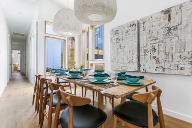 Bright modern home dining room with live-edge wood table set for eight with teal plates and napkins, mid-century wooden chairs, woven pendant lights, large windows and abstract white wall art on light hardwood floors.