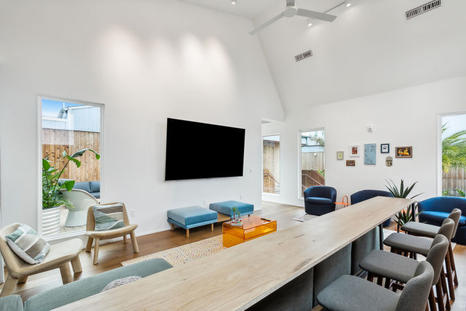 Bright modern living room with vaulted white ceiling, wall-mounted flat-screen TV, long wooden counter with gray bar stools, blue and wood seating, indoor plants, large windows and natural light
