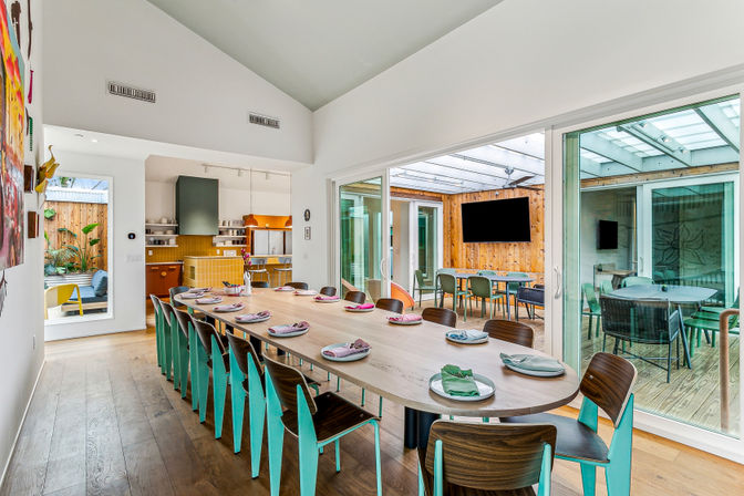 Sunlit modern dining room with a long wooden table surrounded by teal-accent chairs set for a meal, vaulted ceiling and hardwood floors, open kitchen, and sliding glass doors to a covered patio with outdoor seating and wall-mounted TV.