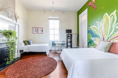 Bright, light-filled bedroom with a large botanical mural on a green accent wall, white linens on a large bed and a smaller bed, hardwood floors with a round rust rug, tall window, small round table with chairs, and a fireplace mantle with a trailing potted plant.