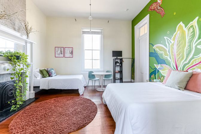 Bright, light-filled bedroom with a large botanical mural on a green accent wall, white linens on a large bed and a smaller bed, hardwood floors with a round rust rug, tall window, small round table with chairs, and a fireplace mantle with a trailing potted plant.
