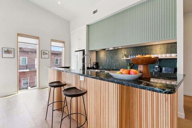 Sleek urban apartment kitchen with black marble breakfast bar, fluted wood island, two black stools, green textured backsplash, fruit bowls and tall windows overlooking brick buildings.