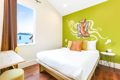 Bright modern bedroom with lime-green accent wall and playful koi fish mural, white bed on a wooden platform with orange and yellow pillows, hardwood floor, small round side tables, desk by a window with brown curtain and neighborhood view.