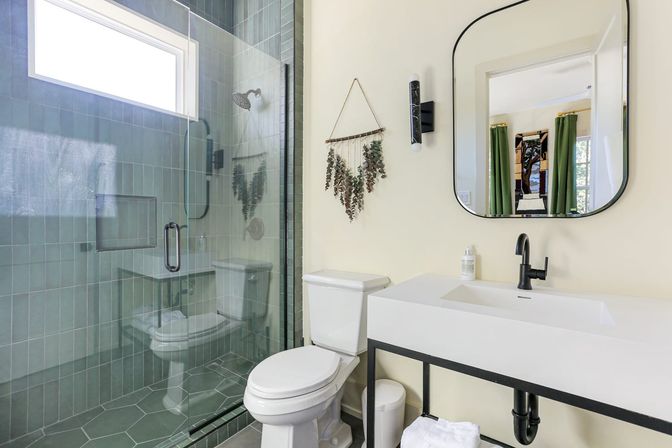 Bright modern bathroom with walk-in glass shower and green tile walls, hex floor tiles, white toilet and minimalist white vanity with black faucet, rounded mirror, hanging dried eucalyptus and natural light.