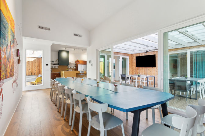 Bright modern communal dining room with a long blue table and white chairs, wood floors, open kitchen, and glass sliding doors opening to a sunlit enclosed patio with a wall-mounted TV.