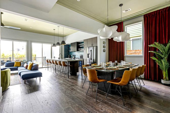 Sunlit modern open-plan living area with dark hardwood floors, a long kitchen island with bar stools, contemporary pendant lights, blue sectional sofa, dining table set with tan leather chairs, bold red curtains and a potted palm — stylish suburban entertaining space.