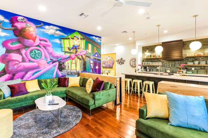 Bright open-plan living room and kitchen with green velvet sectional, hardwood floors, breakfast bar with mint stools and pendant lights, round coffee table, colorful throw pillows and a large vibrant wall mural of a pink character and street scene