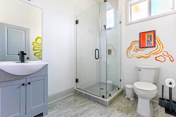 Bright modern residential bathroom with frameless glass corner shower, gray tile floor, light gray vanity with black faucet, white toilet, and playful orange abstract wall art under a small window.