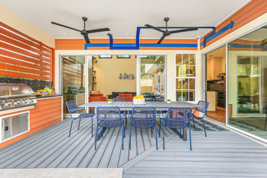 Covered modern backyard patio with long dining table and eight blue metal chairs, built-in stainless steel grill and outdoor kitchen, dual ceiling fans, sliding glass doors to the living room, orange siding and gray composite decking — perfect for al fresco dining.