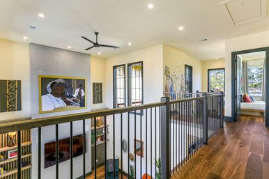 Sunlit upstairs landing in a modern open-concept home with warm hardwood floors, black metal railing overlooking a double-height living room, large framed portrait artwork and mural, ceiling fan, and windows leading to a bright bedroom.