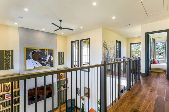 Sunlit upstairs landing in a modern open-concept home with warm hardwood floors, black metal railing overlooking a double-height living room, large framed portrait artwork and mural, ceiling fan, and windows leading to a bright bedroom.