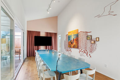 Bright modern dining room with a long turquoise table and white chairs, seascape painting and coral-inspired wall art, floor-to-ceiling sliding glass doors opening to a sunlit deck and warm hardwood floors.