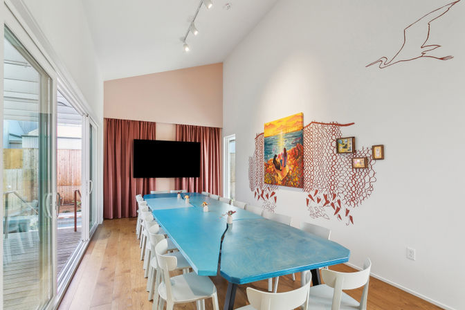 Bright modern dining room with a long turquoise table and white chairs, seascape painting and coral-inspired wall art, floor-to-ceiling sliding glass doors opening to a sunlit deck and warm hardwood floors.