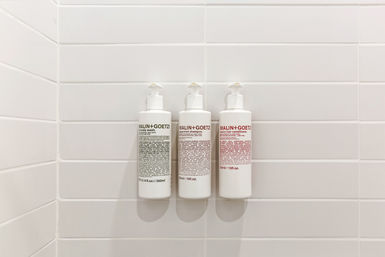 Three white pump dispensers (shampoo, conditioner, body wash) wall-mounted on a white subway-tile shower — minimalist bathroom amenity setup.