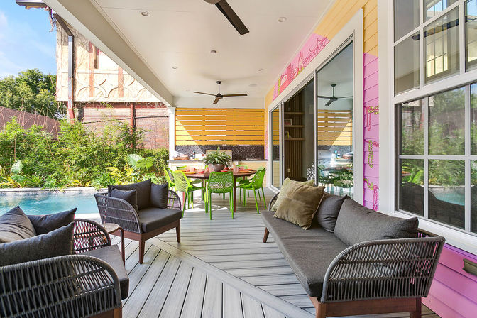 Modern covered poolside patio with gray wicker sofas and cushions, vibrant green dining chairs around a table, sliding glass doors, pink-and-yellow exterior siding, ceiling fans, and lush tropical plants by the pool.