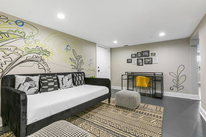 Cozy modern finished basement living area with botanical wall mural, black velvet daybed with patterned throw pillows, geometric rug, knitted pouf, and a small black desk with a yellow accent chair under recessed lighting.