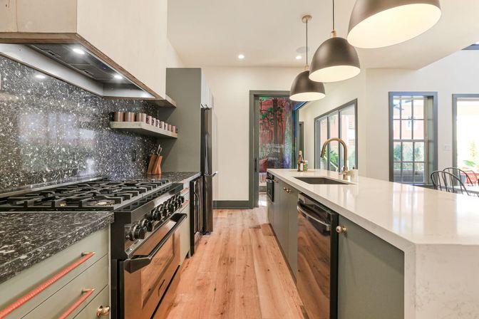Sleek modern kitchen with white quartz island and brass faucet, black stainless range and refrigerator, dark marble countertops and backsplash, three oversized pendant lights, warm hardwood floors and large windows.