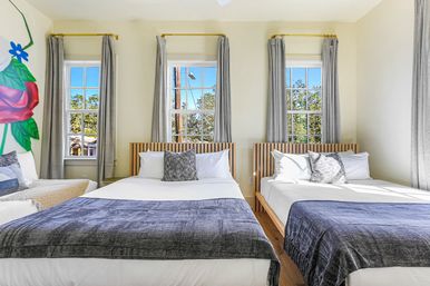 Bright sunlit guest room with two beds, slatted wood headboards, gray curtains framing three tall windows, blue-gray throws and patterned pillows, and a colorful oversized floral mural on the side wall.
