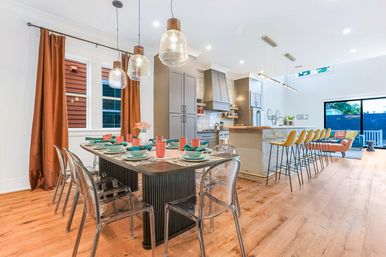 Bright contemporary open-plan kitchen and dining area with hardwood floors, a long table set with teal dishes and clear acrylic chairs, glass pendant lights, a large island with yellow bar stools, and sliding glass doors to a patio.