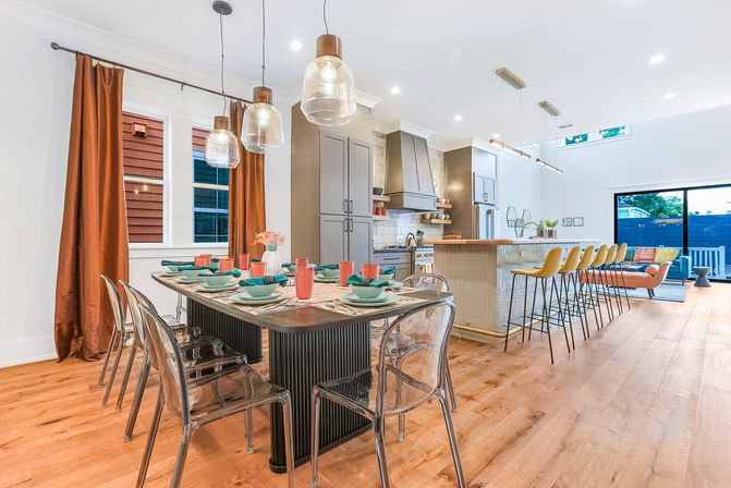 Bright contemporary open-plan kitchen and dining area with hardwood floors, a long table set with teal dishes and clear acrylic chairs, glass pendant lights, a large island with yellow bar stools, and sliding glass doors to a patio.
