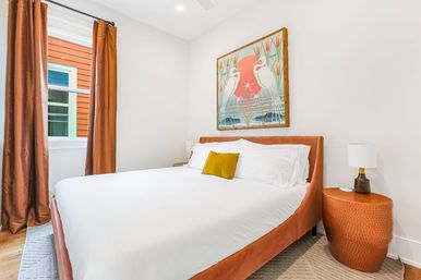 Bright modern bedroom with rust-orange velvet bedframe, crisp white linens and mustard pillow, matching curtains, hammered side table with lamp, bird artwork above bed and sunlit window showing orange exterior siding.