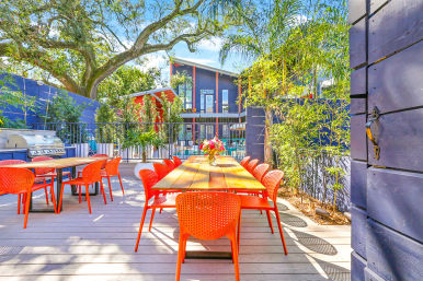 Sunlit backyard patio with long wooden dining table and bright orange chairs on a deck, surrounded by bamboo and a large oak, grill and modern two-story house in the background.