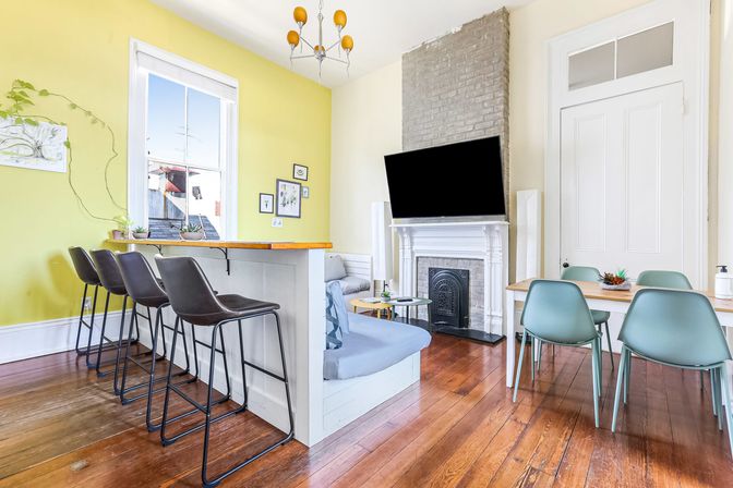 Bright open-plan living room with yellow accent wall, tall window and natural light, wooden breakfast bar with four black bar stools, light-blue built-in banquette, wall-mounted TV above a white fireplace, hardwood floors, dining table with teal chairs, and small plants and framed art.