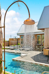 Copper outdoor shower pouring water into a bright blue backyard pool beside a modern wood‑clad house and sunny patio