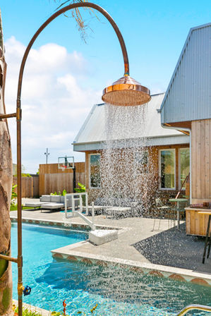 Copper outdoor shower pouring water into a bright blue backyard pool beside a modern wood‑clad house and sunny patio
