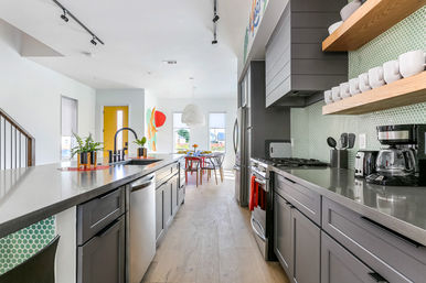 Sunlit modern open-concept kitchen in a suburban home — long island with sink and bar seating, stainless appliances, green penny-tile backsplash, wood shelves, and a yellow entry door.