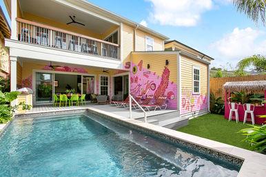 Two-story yellow house backyard with rectangular pool, covered patio and upper balcony, lime-green outdoor dining set, sun loungers, vibrant pink mural on the exterior wall and a tiki-style bar on artificial turf.