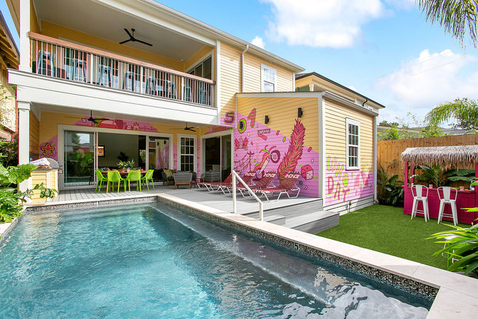 Two-story yellow house backyard with rectangular pool, covered patio and upper balcony, lime-green outdoor dining set, sun loungers, vibrant pink mural on the exterior wall and a tiki-style bar on artificial turf.