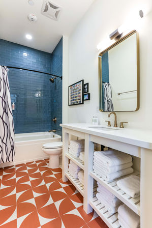 Bright modern bathroom with glossy blue subway-tile shower surround, white tub, brass fixtures and curved black shower rod, terracotta-and-white geometric floor tiles, open white vanity with stacked towels and gold-framed mirror.