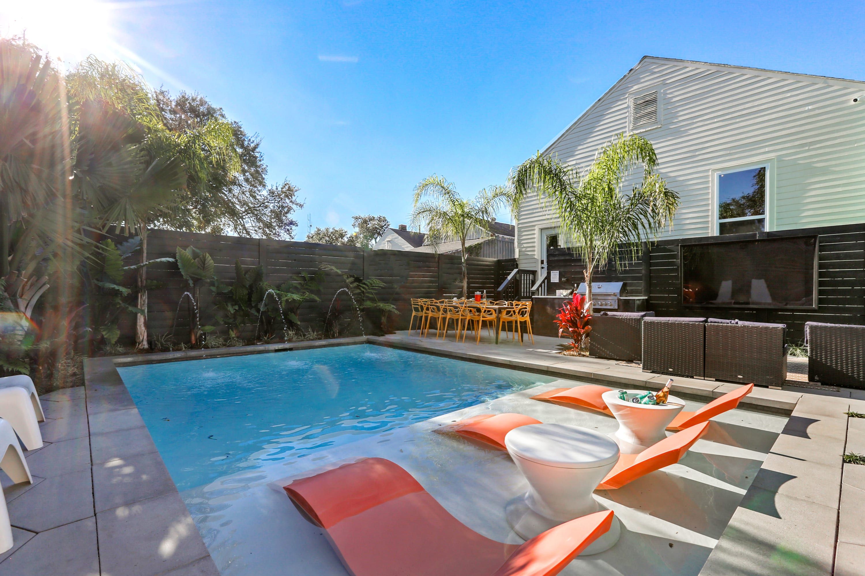 Sunlit backyard pool oasis with a rectangular pool, shallow tanning ledge featuring orange in-water loungers and round white side tables with drinks, palm trees, yellow outdoor dining chairs and wicker lounge seating beside a white house.